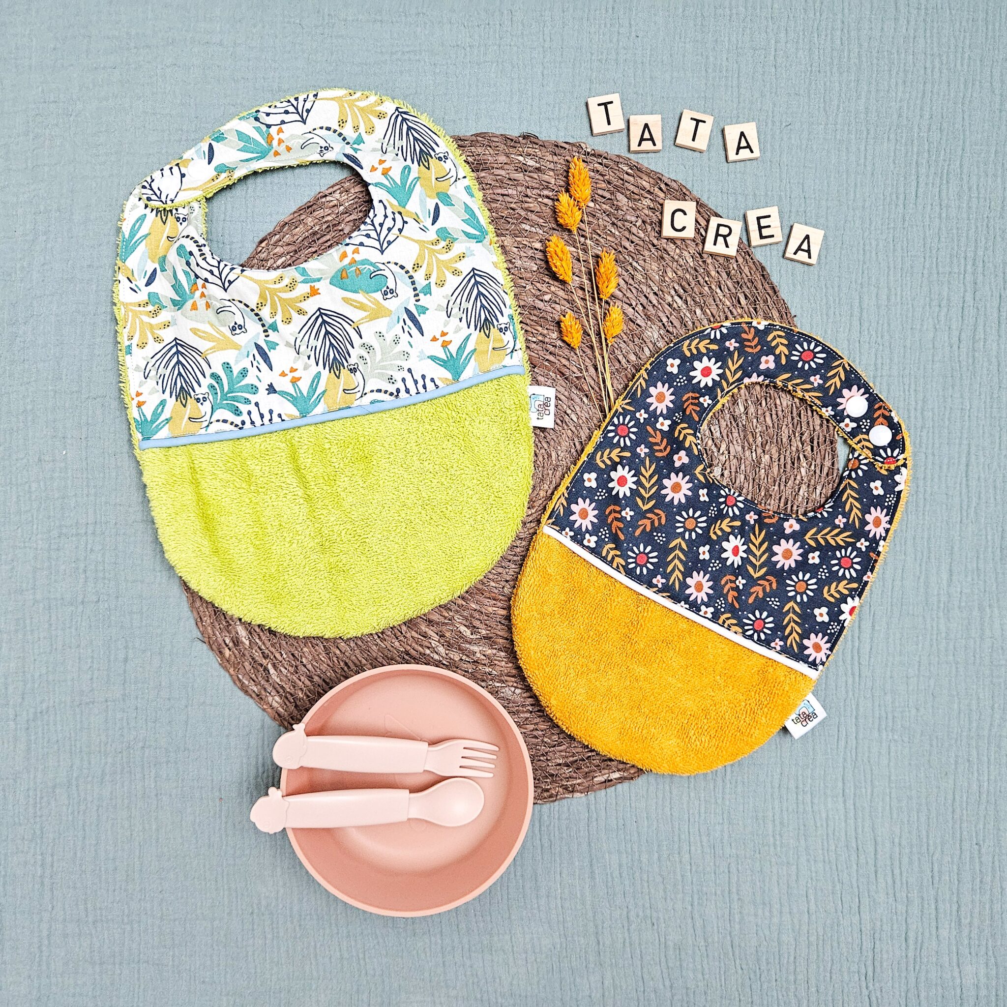 Comparatif de taille entre 2 bavoirs : un naissance (0 à 3 mois) - coton à motifs "fleurs de la prairie", liseré intermédiaire blanc, éponge moutarde // et un bébé (4 mois et plus) - coton à motifs "lémuriens bleus", liseré intermédiaire turquoise, éponge vert anis – modèles Tata Créa
