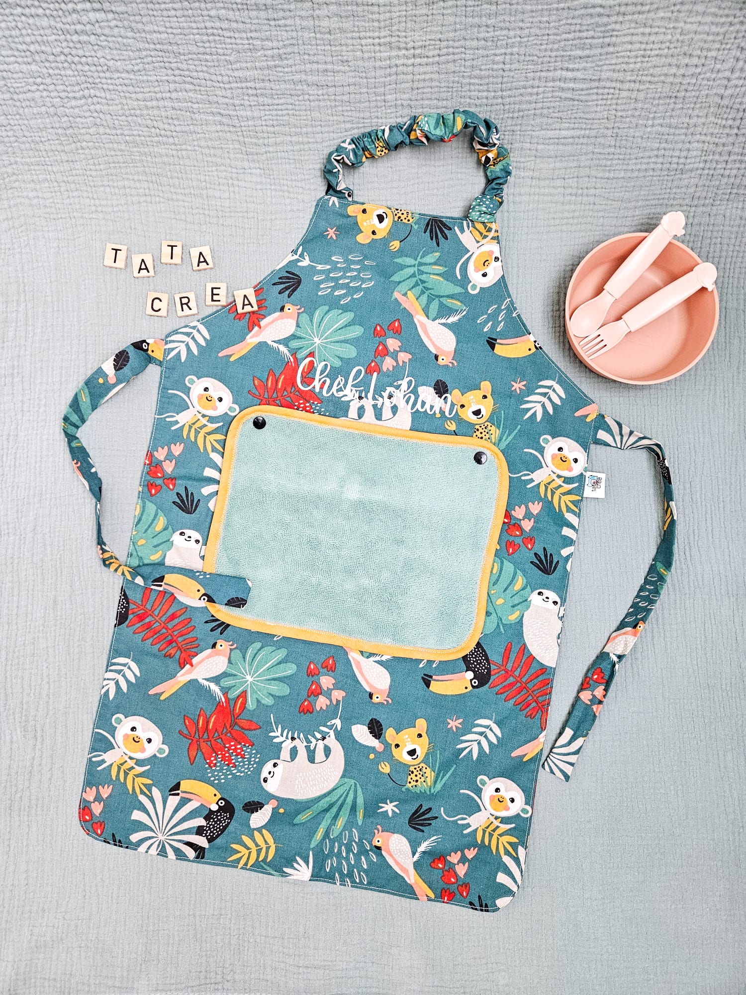 Tablier personnalisé au prénom de Lohan. Tissu motifs "vert singe papaya", avec petite serviette éponge vert tilleul et biais ocre – modèle Tata Créa