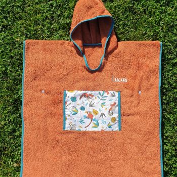 Poncho de bain personnalisé au prénom de Lucas, éponge terra cota et biais bleu pétrole, avec une poche plaquée en coton motifs animaux devant le ventre – modèle Tata Créa